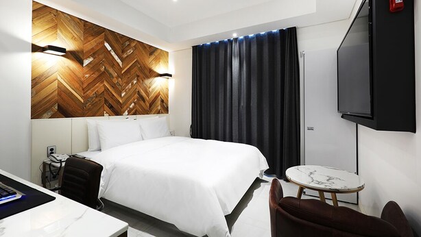 A hotel room with a large bed, a desk with a computer, a small round table, and a TV mounted on the wall. - Alma Business Hotel (Jincheon)