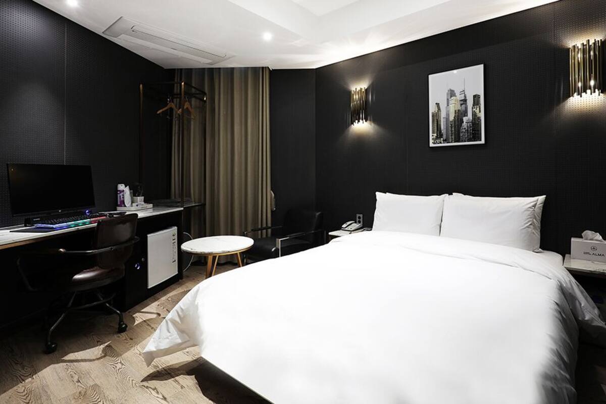A modern hotel room with a large bed, a desk with a computer, a chair, a small round table, and a shower area.