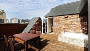 A rooftop terrace with a wooden deck, a brick wall, a small brick building with a door, a table with chairs, and a view of the cityscape. - Alma Business Hotel (Jincheon)