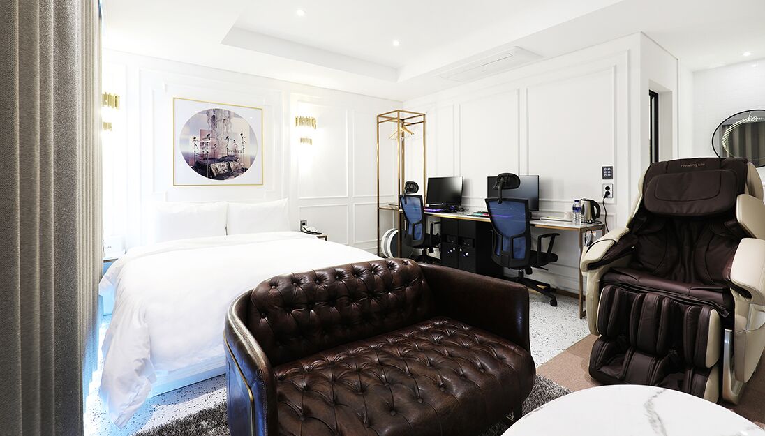A hotel room with a large bed, a massage chair, a desk with a computer, and a massage table.