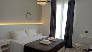 Ein ordentlich bezogenes Bett mit weißen Leinentüchern, einer dunkelbraunen Tagesdecke und zwei gefalteten Handtüchern am Fußende. Ein kleiner Nachttisch mit einer Lampe und einem an der Wand befestigten dekorativen Gegenstand sind ebenfalls zu sehen.