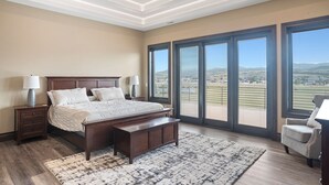 Ein Schlafzimmer mit einem großen Bett, einem Nachttisch, einer Bank und einem Sessel. Große Fenster bieten Blick auf eine entfernte Landschaft.