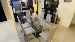 A modern dining area with a glass table and six chairs, a kitchen in the background, and a decorative wall piece. - Luxurious Home (Draxhall)