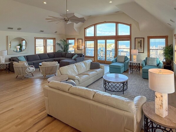 A spacious living room with a large sectional sofa, a chaise lounge, and a round coffee table. There are multiple seating options including a small round table and a couple of chairs. The room features large windows with a view of the outdoors and a ceiling fan.