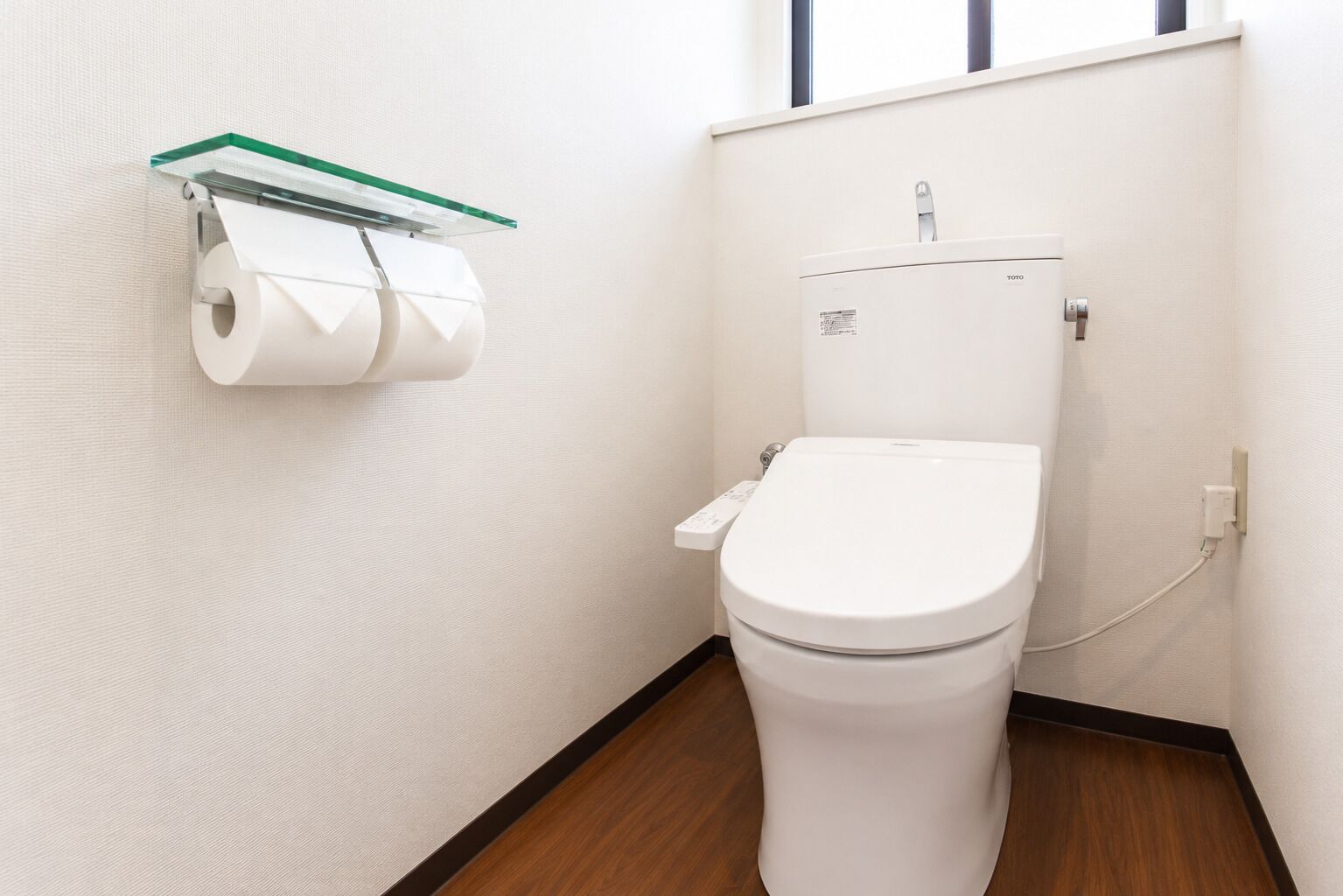 A toilet with a Toto brand label, a glass shelf with toilet paper, and a wooden floor.