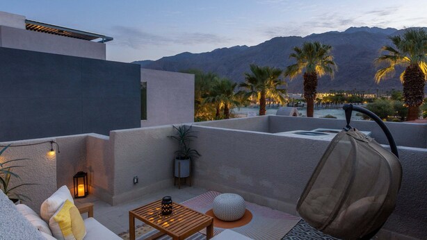 A rooftop terrace with a sofa, table, and chairs, overlooking a mountainous landscape with palm trees.