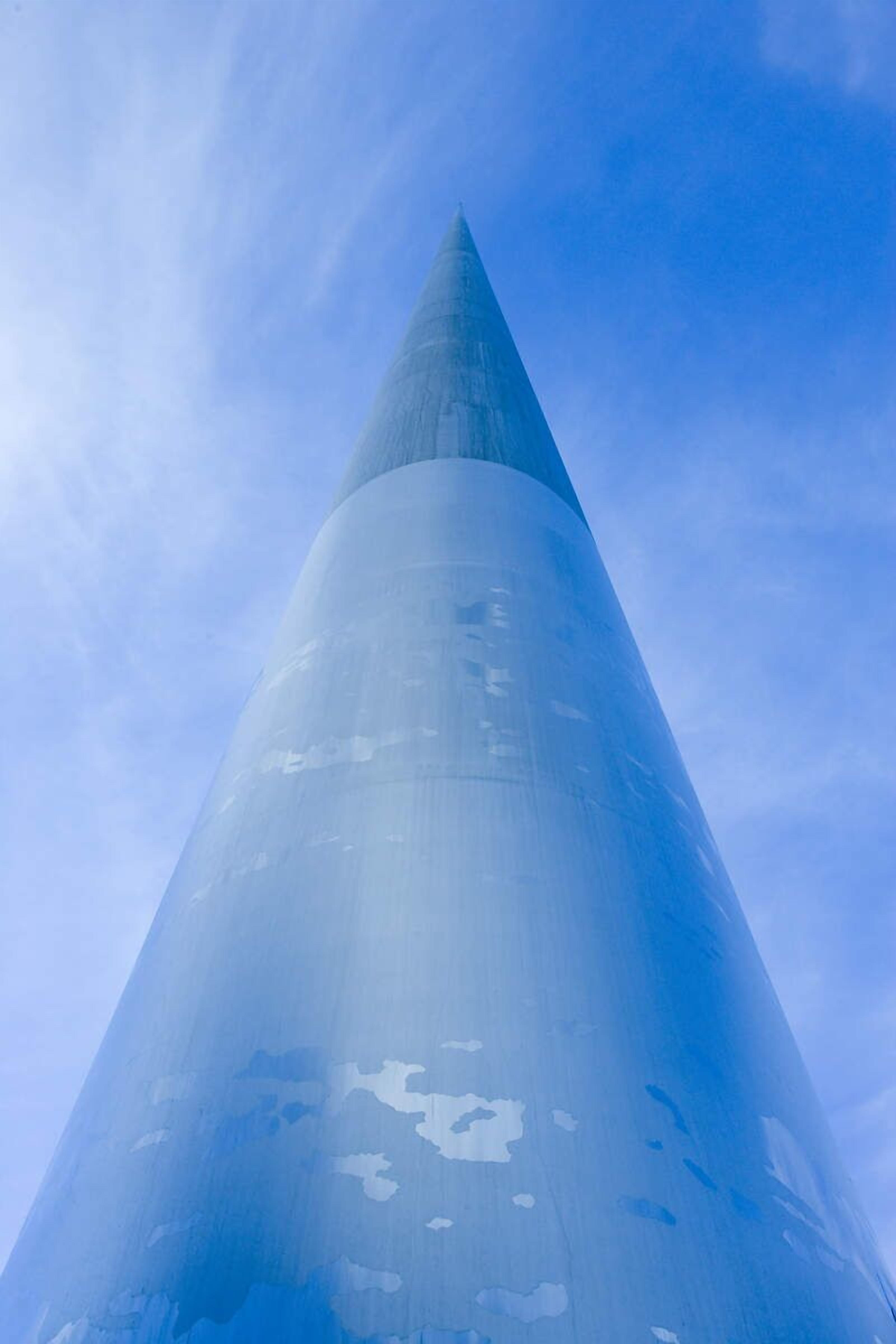 Une structure conique se terminant par un sommet pointu, qui pourrait être un monument ou une sculpture, se détachant sur un ciel dégagé.