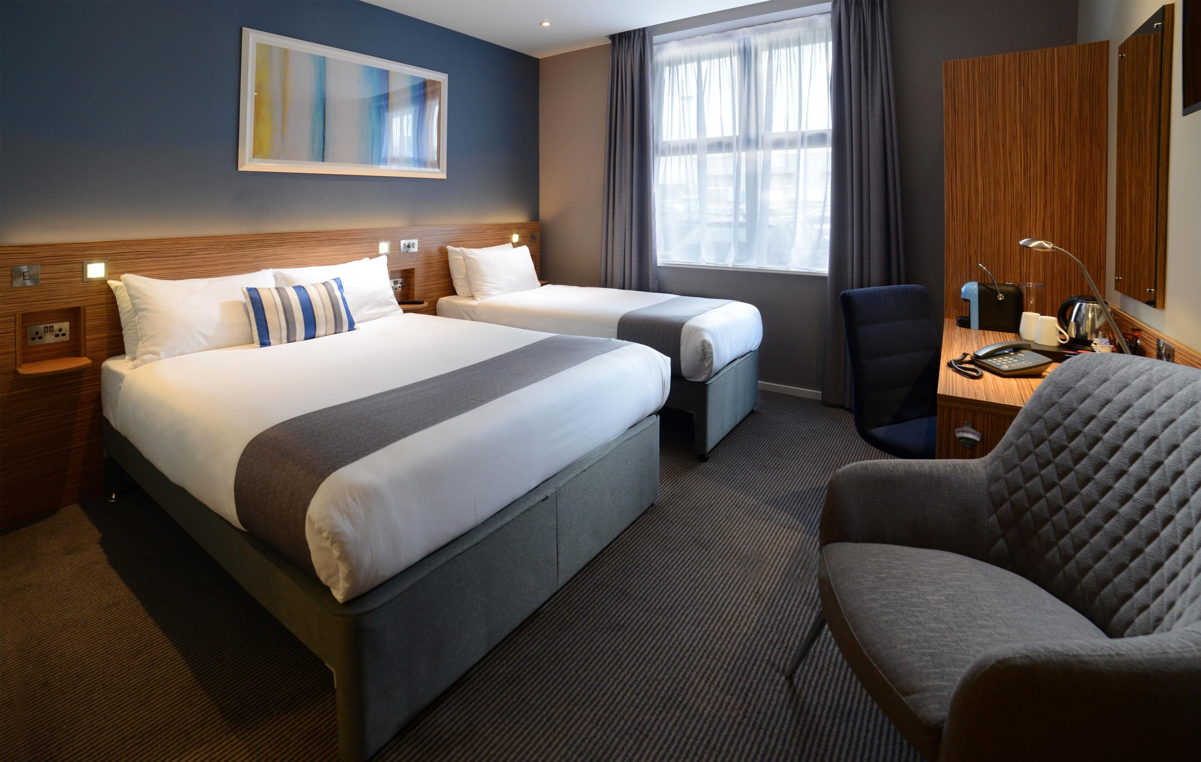 A hotel room with two beds, a desk, a chair, a lamp, and a large window with curtains.