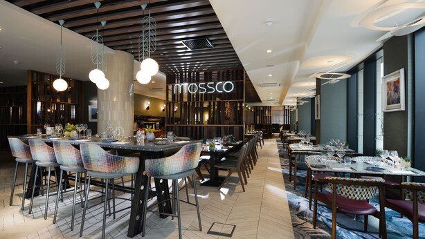 Un interior de restaurante moderno con varias mesas y sillas, un letrero visible de 'Mossco' y una combinación de diseños de techo.