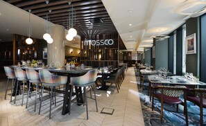 Een modern restaurantinterieur met meerdere eettafels en stoelen, een zichtbaar 'Mossco'-bord en verschillende soorten plafondontwerpen.