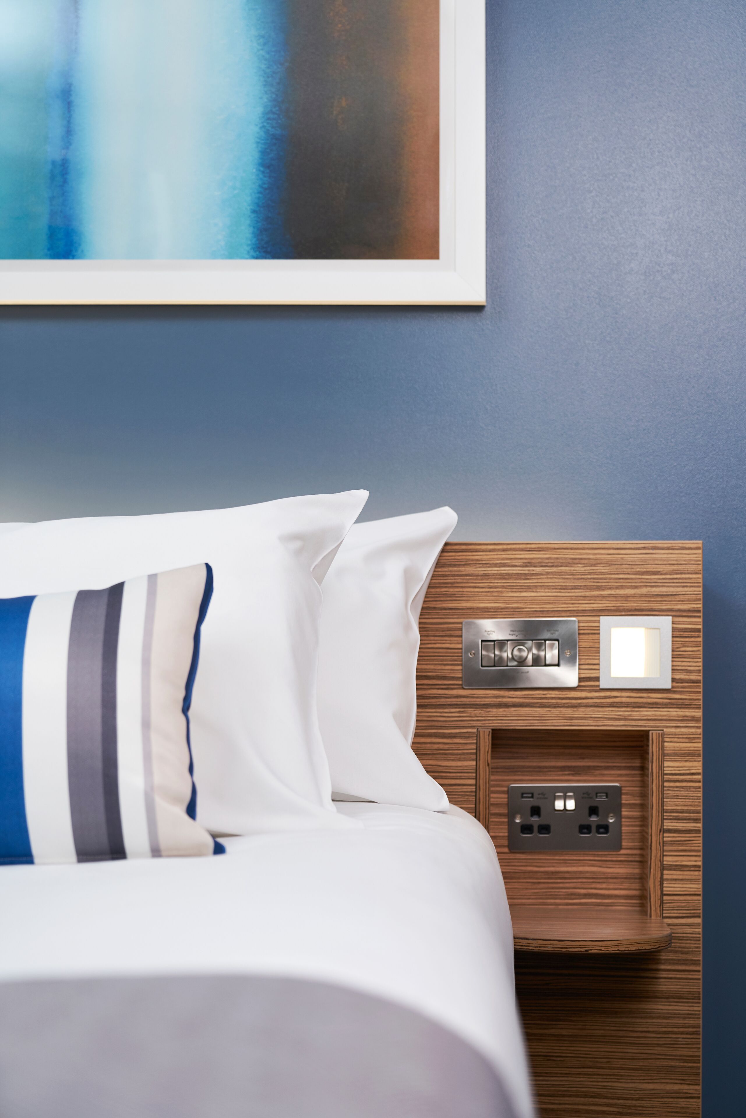 Una cama bien hecha con almohadas blancas y a rayas, un cabecero de madera con toma de corriente y un cuadro abstracto enmarcado en la pared.