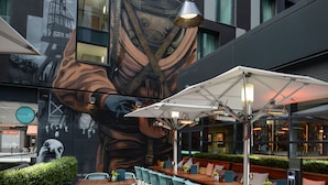 Un espacio al aire libre con mesas y sillas, un mural en el edificio y sombrillas que dan sombra.