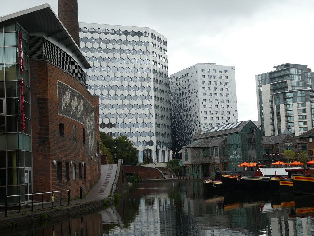 Un edificio moderno con una fachada hexagonal distintiva, un canal con barcos y un edificio tradicional con un letrero que dice "THE CANAL HOUSE".