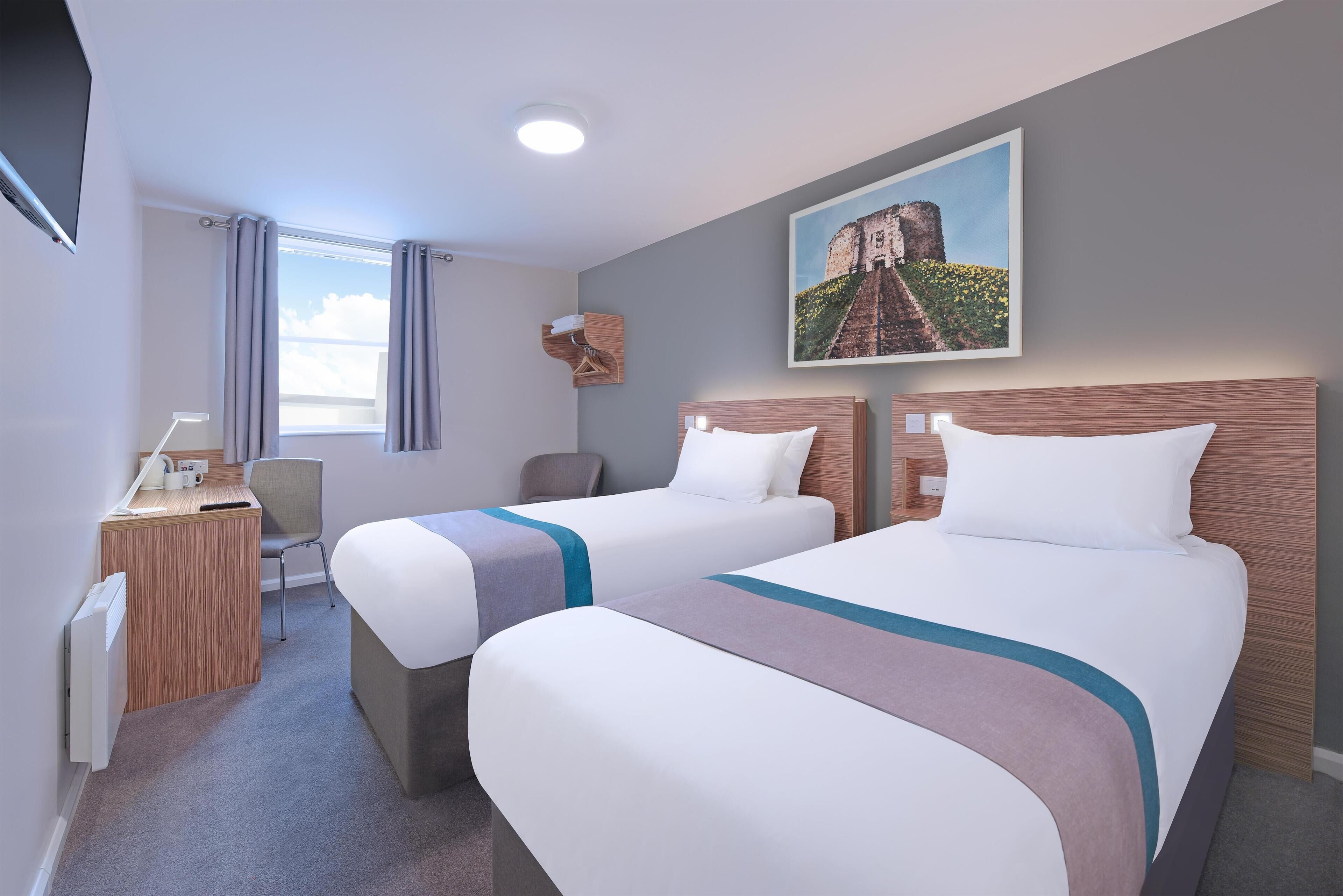 A hotel room with two beds, a desk, a chair, a window with curtains, and a painting on the wall.