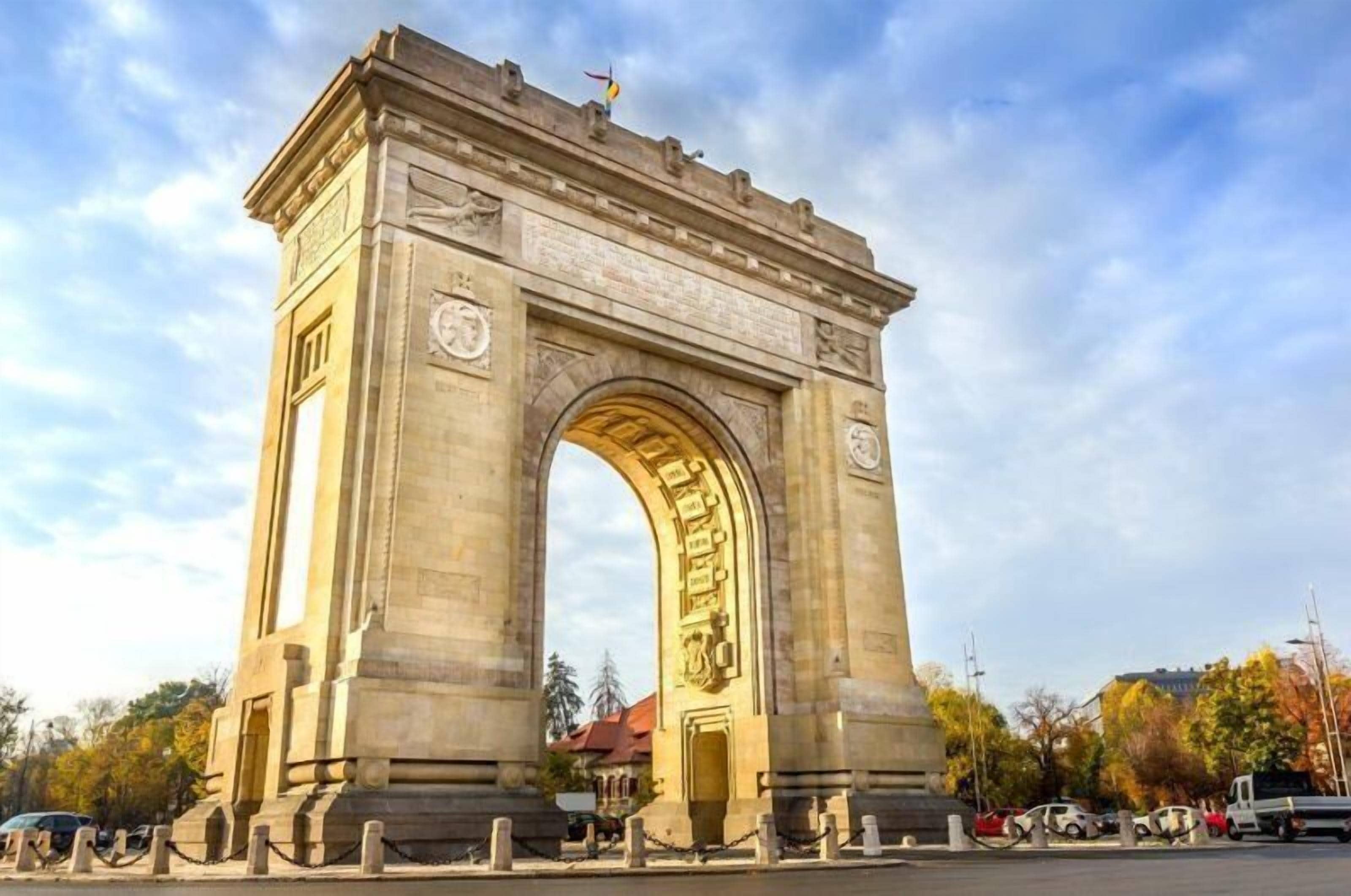 A large stone arch monument with inscriptions, surrounded by a chain-link fence and parked cars.