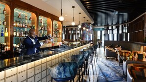 Una zona de bar con un bartender preparando bebidas, una variedad de botellas en estantes y asientos con alfombra estampada.