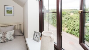 A bedroom with a large window, a framed picture on the wall, and a bedside table with a vase. - Blueberry Cottage (Dorchester)