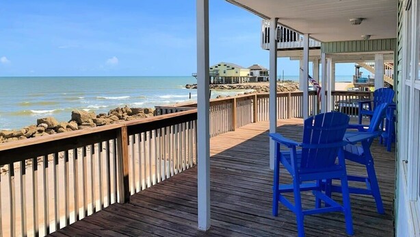 Een houten terras met blauwe stoelen met uitzicht op de oceaan, een houten reling, en een beeld van het strand en de zee.