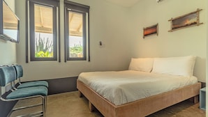 Un dormitorio con una cama, dos ventanas, una silla y un televisor instalado en la pared.