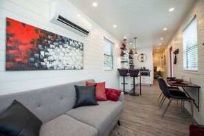 Ein modernes Wohnzimmer mit einem grauen Sofa, einem abstraktem Gemälde in Rot und Schwarz und einer Küchenzeile mit Esstisch und Stühlen.