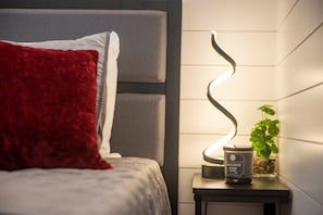 A bed with a red pillow, a white pillow, and a grey headboard. There is a small table with a plant and a candle. - The Merlot - Unique Container Home Near Waco Attra (Waco)