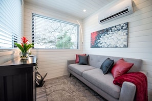 A living room with a grey sofa, red and black cushions, a small table with a plant, and a painting on the wall. - The Merlot - Unique Container Home Near Waco Attra (Waco)
