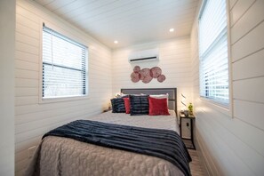 A bedroom with a bed, a nightstand, a wall-mounted air conditioner, and a window with blinds. - The Merlot - Unique Container Home Near Waco Attra (Waco)
