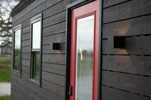 A building with a red door, two windows, and a small square window on the wall. - The Merlot - Unique Container Home Near Waco Attra (Waco)