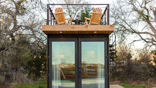 A modern cabin with a deck, two wooden chairs, and a glass door. - Deer Creek Tiny Container Home-12 Min to Magnolia (Waco)