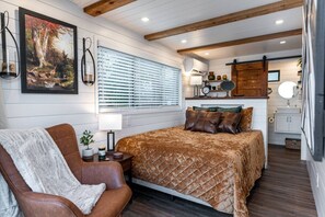 Une chambre à coucher comprenant un lit, une chaise, une table de chevet, une fenêtre avec des stores, un tableau et un plafond en bois.