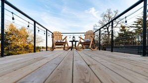 A wooden deck with two rocking chairs and a table, surrounded by metal railings and string lights. - Deer Creek Tiny Container Home-12 Min to Magnolia (Waco)