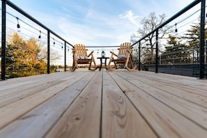 En træterrasse med to gyngestole og et bord, omgivet af gelændere og lyskæder.
