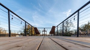 A wooden deck with two wooden chairs and a table, surrounded by metal railings and trees.
