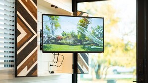 A flat-screen TV mounted on a wall with a geometric pattern, positioned in front of a window with blinds.