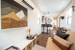 A modern interior with a white paneled wall, a large abstract painting, a sofa with cushions, a dining area with a table and chairs, and a kitchen in the background. - The Newtonian Tiny Container Home near Magnolia (Waco)