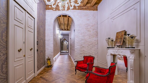 A hallway with wooden parquet flooring, a red upholstered chair, a white fireplace with a mirror, and a door with a brass handle. - Corte Rubbi 5 Deluxe Studio-Apartment (Venice)