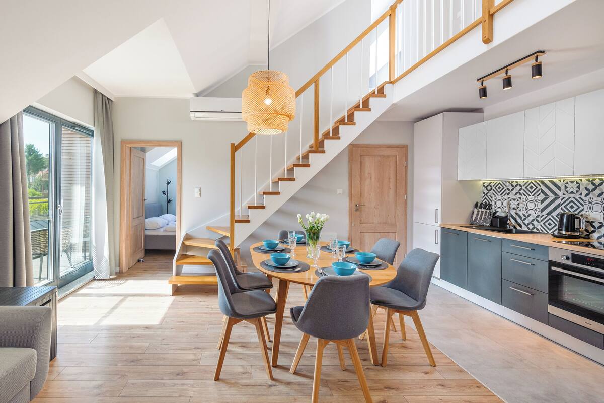 A modern kitchen with a dining area, wooden staircase, and large windows.