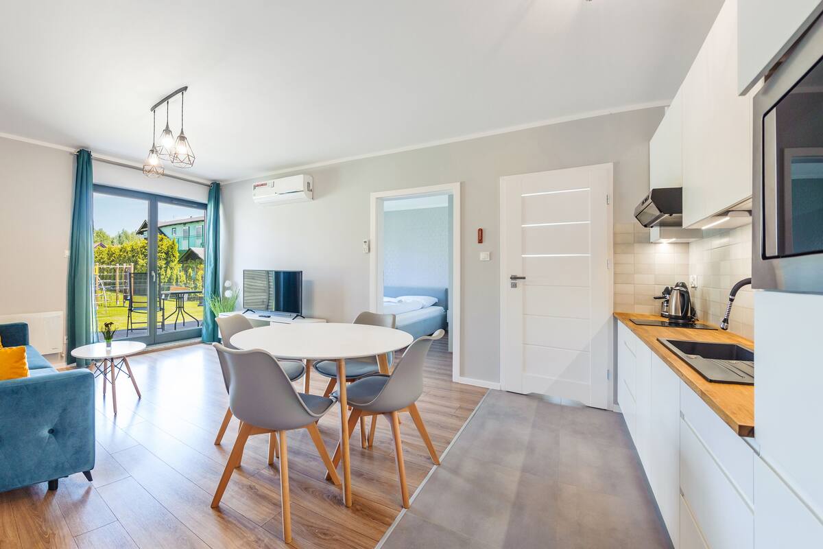 A modern living room with a sofa, a dining table with chairs, a TV, and a large sliding glass door leading to a balcony.