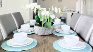 Un comedor con una mesa redonda dispuesta para seis personas, rodeada de sillas tapizadas en gris. Un macetero de metal rústico con orquídeas blancas se encuentra en el centro.
