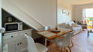 Un coin repas avec une table en bois et des chaises blanches, un canapé blanc et un espace cuisine équipé d’un four à micro-ondes.
