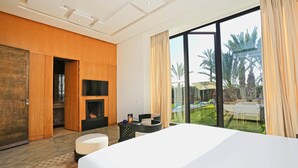 A modern hotel room with a large bed, a fireplace, a TV, and a view of a pool and palm trees. - Impeccable 5-bed Villa in Marrakech (Marrakech)