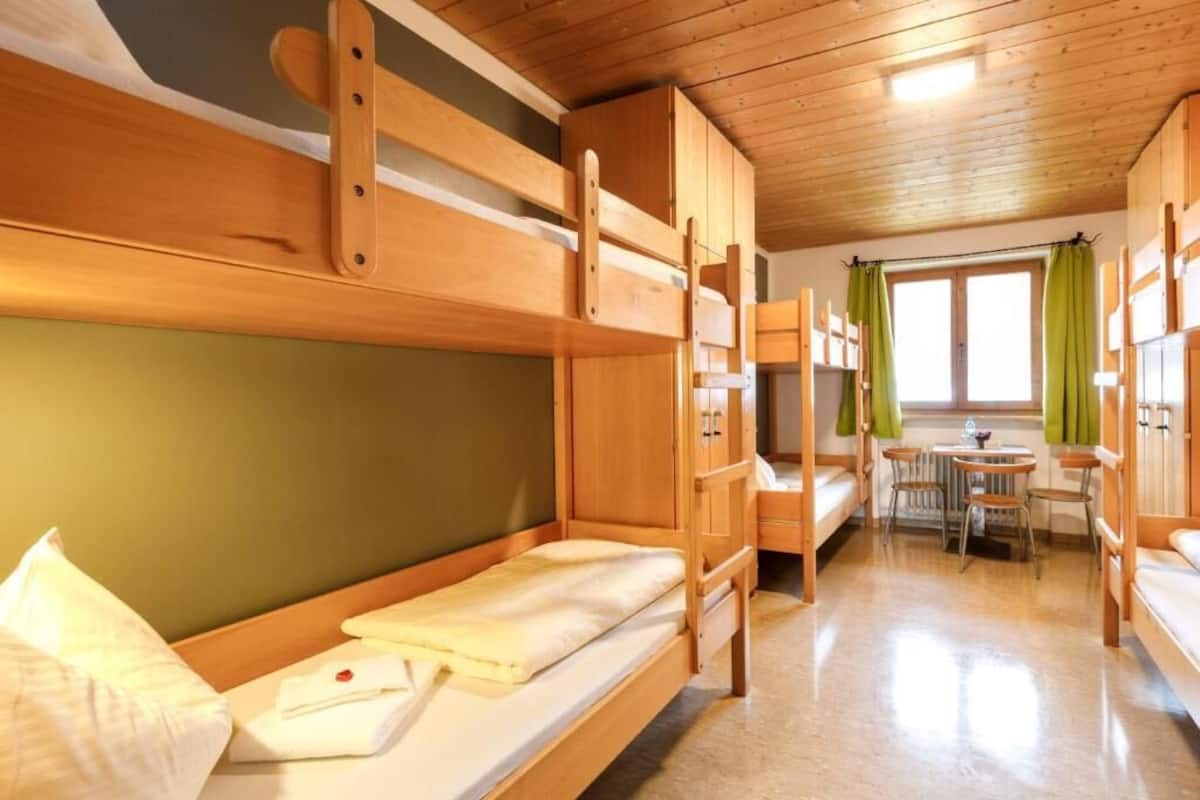 Ein Schlafraum mit Etagenbetten, Holzboden, einem kleinen Tisch mit Stühlen und einem Fenster mit Vorhängen.