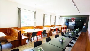 A dining area with wooden benches and tables, large windows with curtains, and a kitchen area visible in the background. - Jugendherberge Kreuth-Scharling - Hostel (Kreuth)