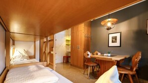 A compact hotel room with a bed, a small dining table, and a chair. There is a wardrobe and a wall-mounted light fixture.