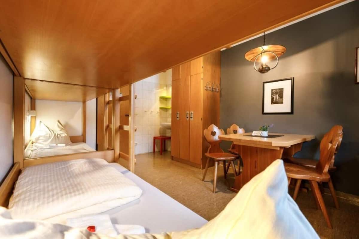 Ein kleines Hotelzimmer mit einem Bett, einem Esstisch und einem Stuhl. Es gibt einen Kleiderschrank und eine Wandleuchte.