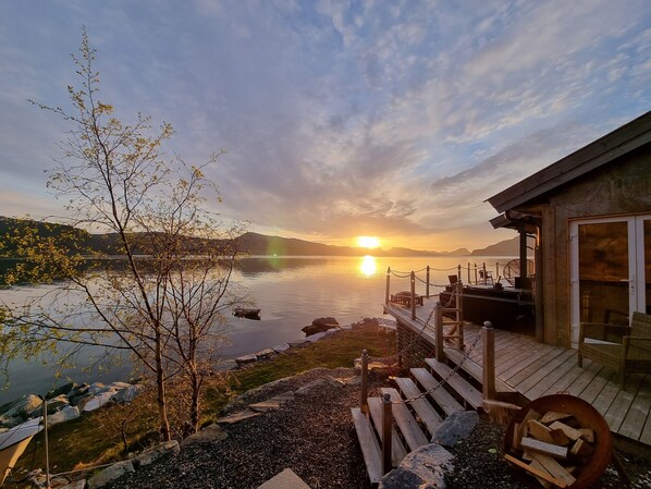 A lakeside cabin with a wooden deck, a fire pit, and a view of the sunset over the water.