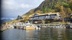 A lakeside cabin with a thatched roof, a dock with boats, and a mountain backdrop.