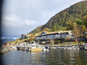 A lakeside cabin with a thatched roof, a dock with boats, and a mountain backdrop. - Nytt Stort Feriehus med Panoramautsikt. Båt Inkludert (Åheim)