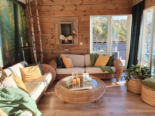 En mysig stuga med träpanel på insidan, en soffa, ett soffbord i rotting och stora fönster med utsikt över naturen.