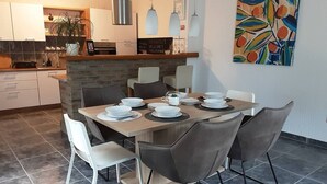 A modern dining area with a wooden table set for six, surrounded by grey chairs. A kitchen with white cabinets and a brick accent wall is visible in the background. - Villa Sorella Beautiful 3-bedroom Villa in Karanac (Knezevi Vinogradi)